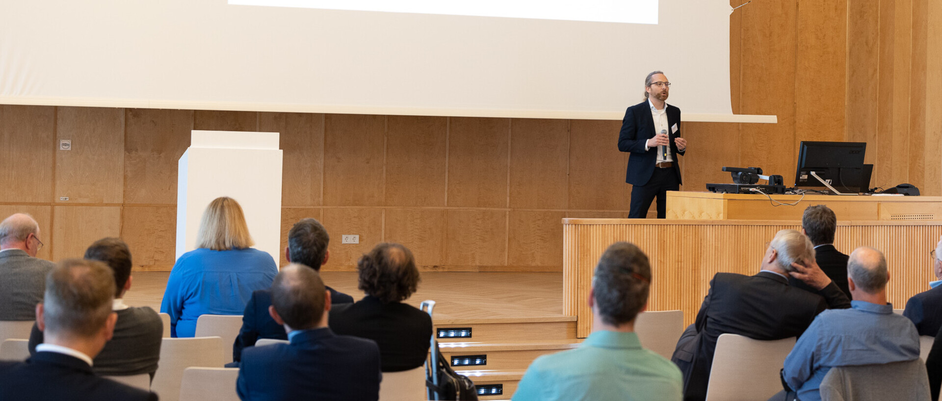 Prof. Heiner Stuckenschmidt in dunklem Anzug und weißem Hemd auf der Bühne der Aula, er hält eine Präsentation, die im Hintergrund zu sehen ist. Im Vordergrund sieht man die ersten Reihen eines Publikums, alle formell gekleidet.