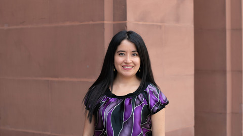 Tania Guerra Rosero. She is wearing a purple-black T-shirt and is standing in front of brown wall.