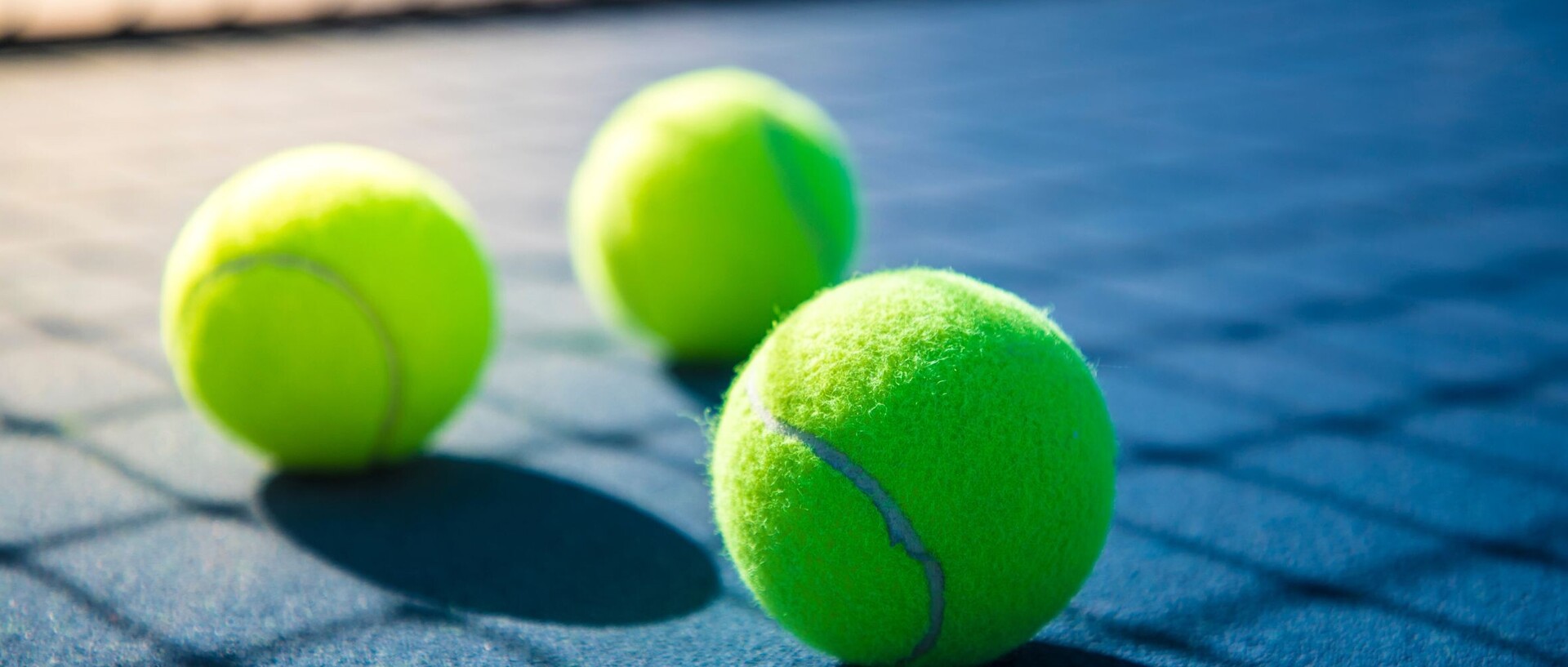 3 gelbe Tennisbälle liegen auf einem Tennisplatz, das Netz wirft einen Schatten auf den Boden