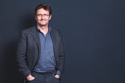 Portrait von Peter Vorderer. Er trägt ein blaues Hemd sowie einen grauen Blazer und steht vor einer dunkelblauen Wand.