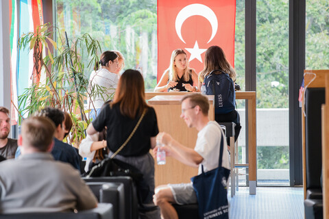 Studierende in der Mensa unterhalten sich, im Hintergrund ist groß die türkische Landesflagge zu sehen.