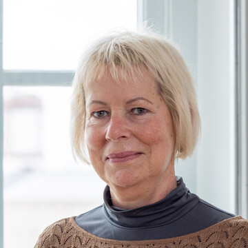 Ute Becker hat blonde mittellange Haare. Sie trägt ein schwarzes Oberteil mit einem baunen Strickpullover darüber.