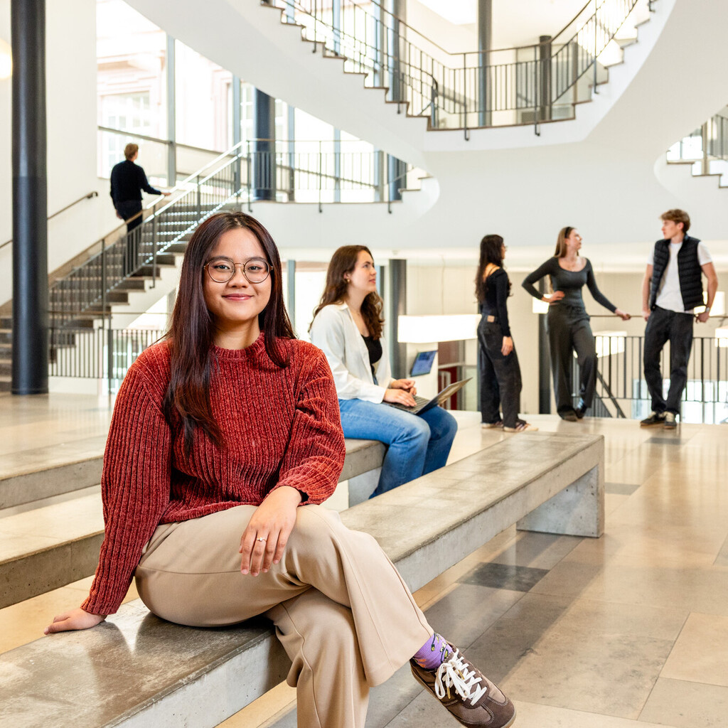 Eine Studentin sitzt auf einer Seitenbank im Ostflügel des Schlosses. Im Hintergund unterhalten sich weitere Studierende. 