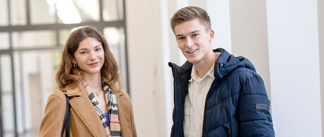 Eine Studentin und ein Student stehen im Flur des Schlosses. Sie tragen Jacken und Laptops unter dem Arm. Sie schauen interessiert in die Kamera.