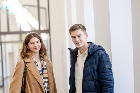 Eine Studentin und ein Student stehen im Flur des Schlosses. Sie tragen Jacken und Laptops unter dem Arm. Sie schauen interessiert in die Kamera.