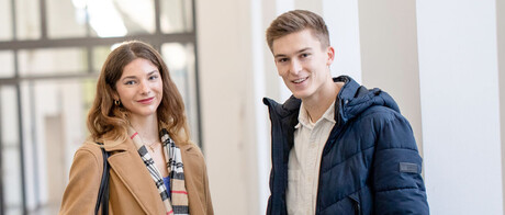 Eine Studentin und ein Student stehen im Flur des Schlosses. Sie tragen Jacken und Laptops unter dem Arm. Sie schauen interessiert in die Kamera.