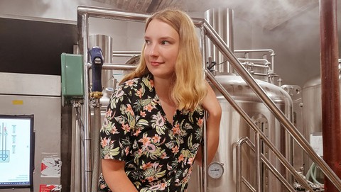 Eine Person trägt eine schwarze Bluse mit Blumen und Früchten und sitzt auf einer Stufe vor einem Kessel einer Bierbrauerei. Die Person heißt Sarah Fent.