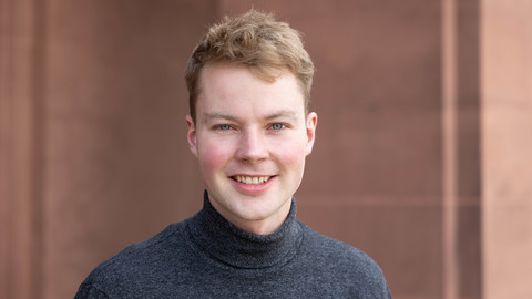 Daniel Linnenbrink. He is wearing a grey sweater and is standing in front of a brown wall.