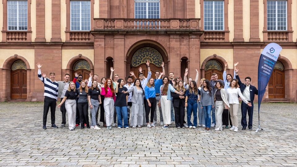Auf dem Ehrenhof steht eine Gruppe von Menschen, die ihre Arme in die Luft strecken. Rechts neben ihnen steht ein Banner von engage.eu.