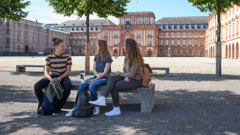Drei Studierende sitzen nebeneinander auf einer Bank auf dem Ehrenhof. Hinter ihnen sind drei Bäume und das Schloss Ehrenhof. 