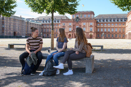 Drei Studierende sitzen nebeneinander auf einer Bank auf dem Ehrenhof. Hinter ihnen sind drei Bäume und das Schloss Ehrenhof. 