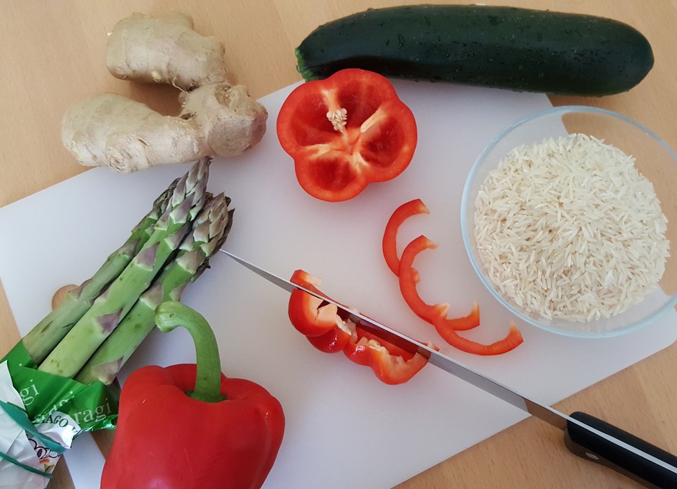 Auf einem Schneidebrett liegt Spargel, Paprika, Zucchini, Ingwer und daneben steht eine Schüssel mit Reis.