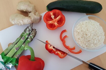 Auf einem Schneidebrett liegt Spargel, Paprika, Zucchini, Ingwer und daneben steht eine Schüssel mit Reis.