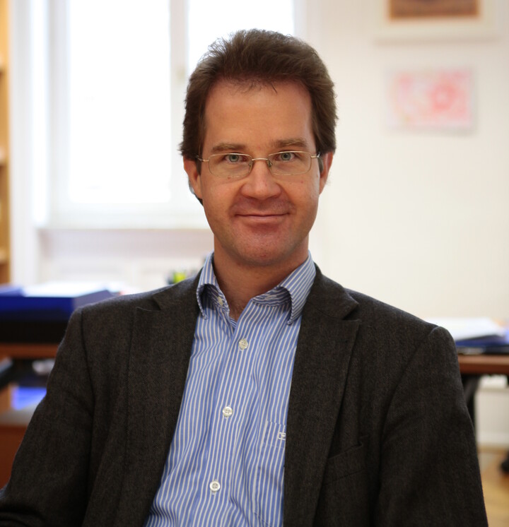 Prof. Dr. Christian Mann mit kurzen braunen Haare, einer eckigen Brille in einem blau-weiß-gestreiftem Hemd und anthrazit-farbenen Sakko in einem Büro
