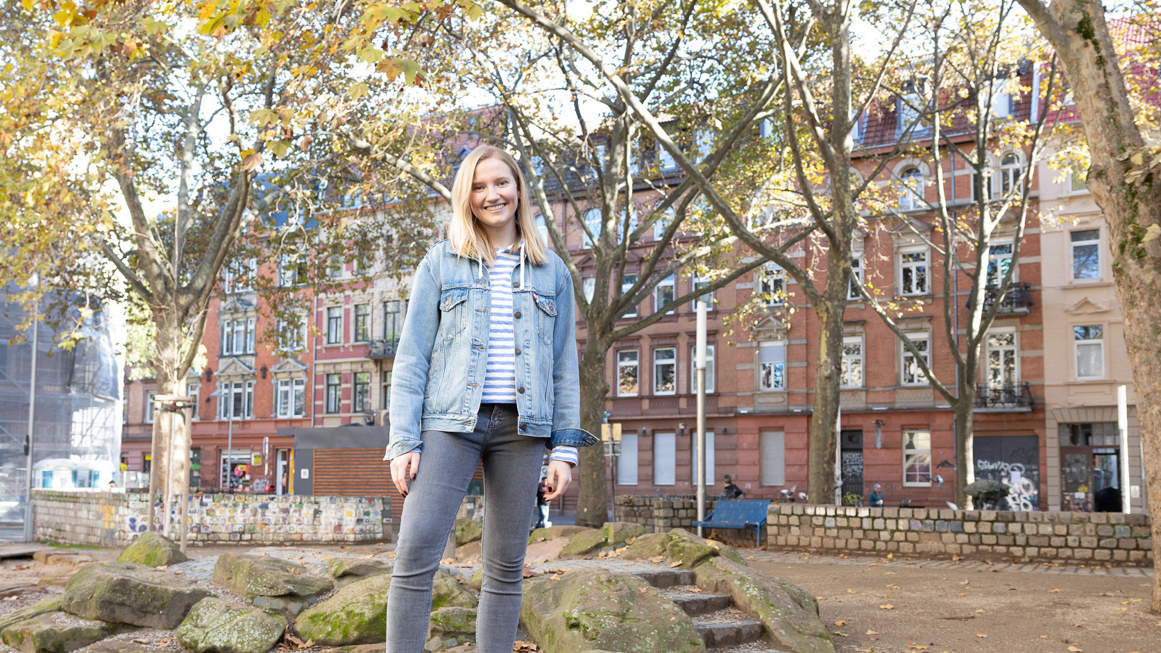 Eine Studentin mit schulterlangen blonden Haaren und einer Jeansjacke steht auf einem Platz im Jungbusch