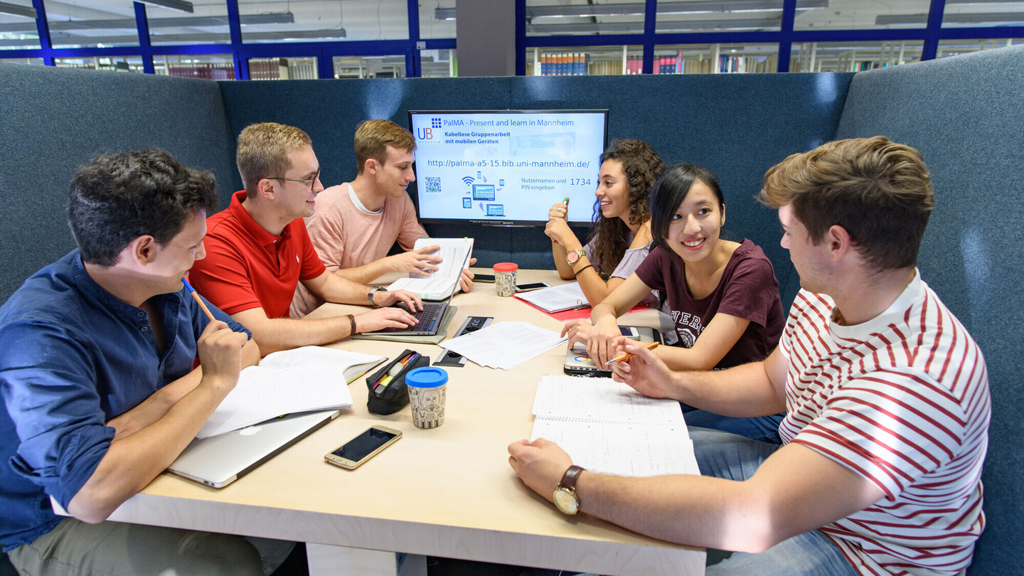 Eine Lerngruppe sitzt um einen Tisch, im Hintergrund ist ein Monitor zu sehen.