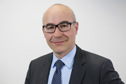 Portrait von Achim Wambach. Er trägt ein hellblaues Hemd sowie eine dunkelblau karrierte Krawatte und einen schwarzen Blazer. Er steht lächelnd vor einer grauen Wand.