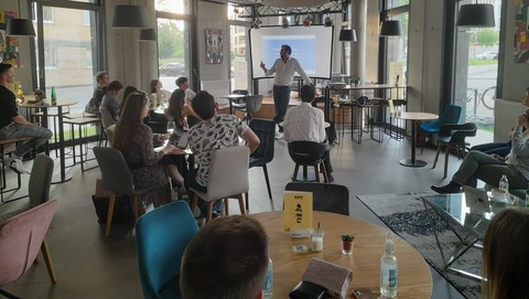 In einem Café sitzen viele Personen. Eine Person mit weißem Hemd steht vor einer Leinwand und spricht.