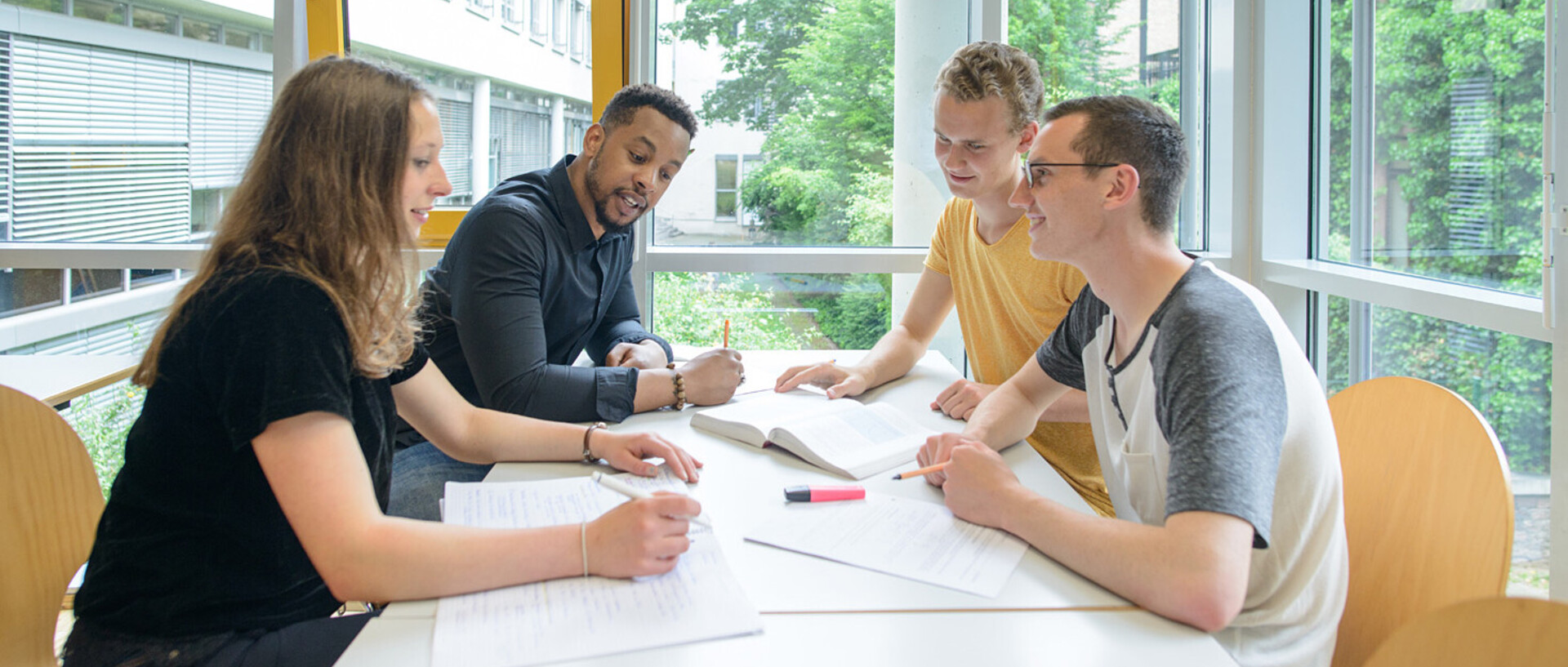 Vier Studierende sitzen in einem freundlichen und hellen Unigebäude und lernen an einem Tisch zusammen