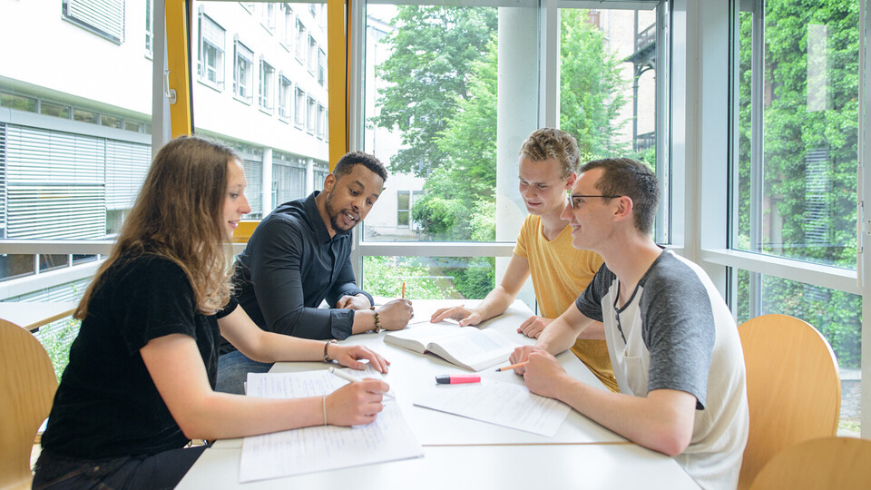 Vier Studierende sitzen in einem freundlichen und hellen Unigebäude und lernen an einem Tisch zusammen