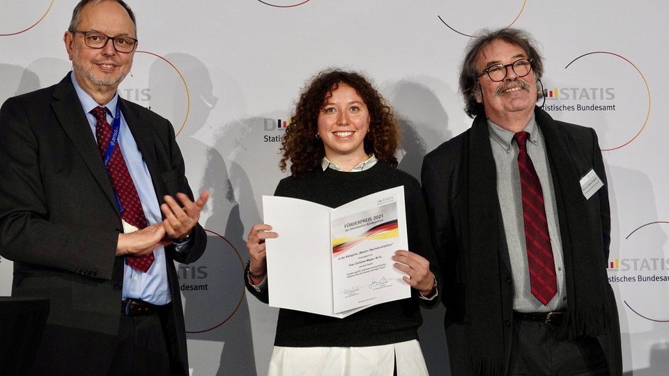 Christina Meyer and two men are standing in front of a white background. She is holding a paper.