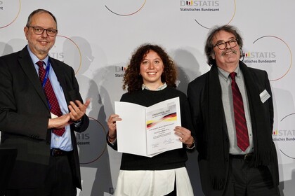 Christina Meyer and two men are standing in front of a white background. She is holding a paper.
