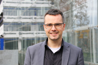 Portrait von Marc Debus. Er trägt ein schwarzes Hemd sowie einen grauen Blazer und steht vor dem A5-Gebäude.