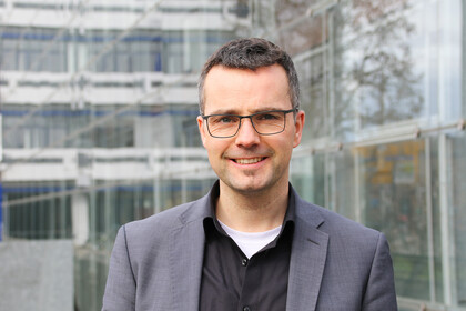 Portrait von Marc Debus. Er trägt ein schwarzes Hemd sowie einen grauen Blazer und steht vor dem A5-Gebäude.