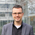 Portrait von Marc Debus. Er trägt ein schwarzes Hemd sowie einen grauen Blazer und steht vor dem A5-Gebäude.