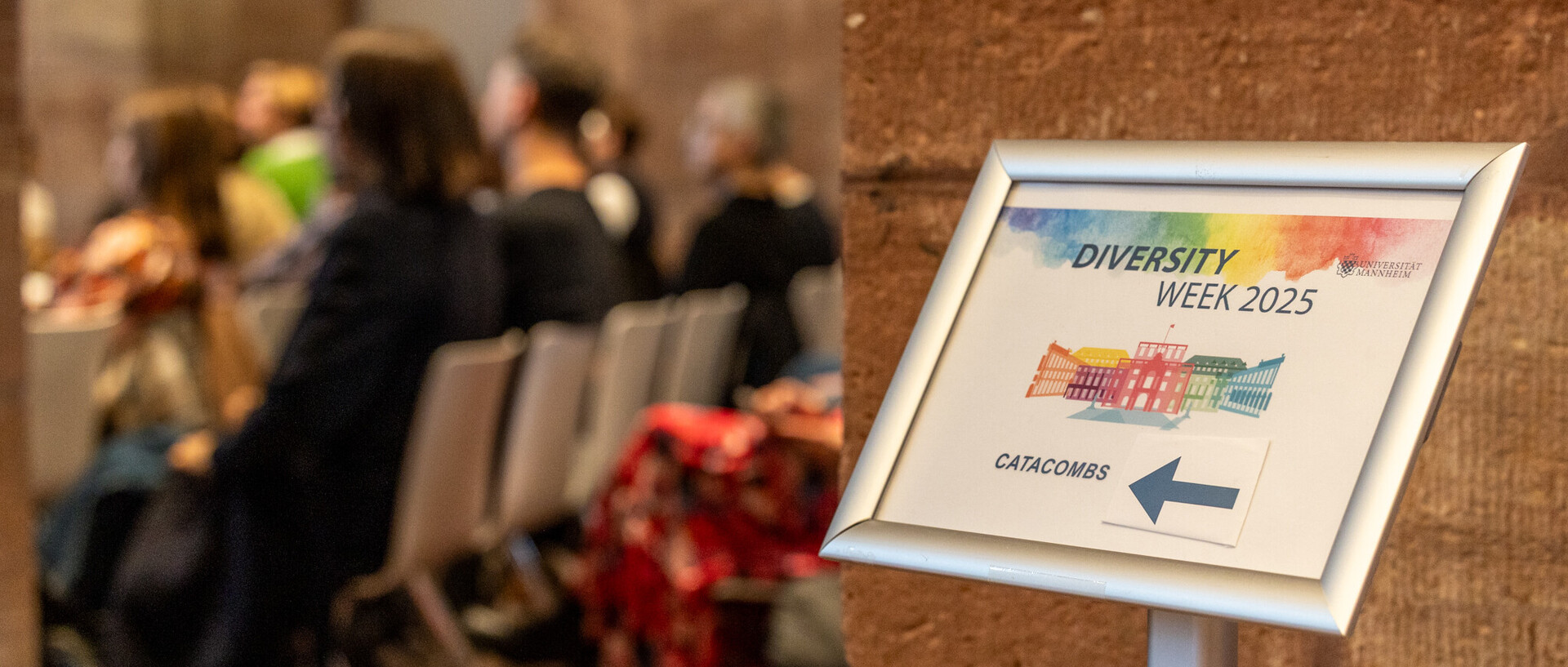 Links: Zuschauer sitzen auf Stühlen im Publikum und sind nur leicht unscharf zu erkennen. Im Vordergrund ist ein Wegweiser mit der Aufschrift "Diversity Week" zu erkennen.
