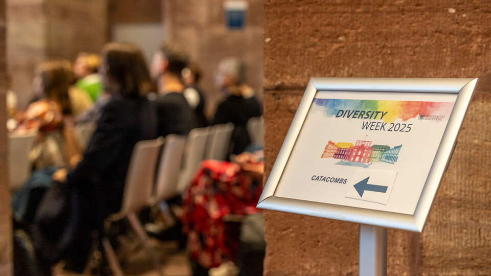 links, Zuschauer sitzen auf Stühlen im Publikum, leicht unscharf. Im Vordergrund ist ein Wegweiser mit der Aufschrift "Diversity Week" zu erkennen.