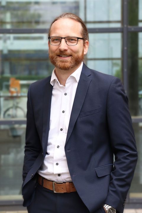 Heiner Stuckenschmidt hat rötlich-braune, zu einem Zopf gebundene Haare. Er trägt eine eckige Brille, ein weißes Hemd und ein dunkles Jacket