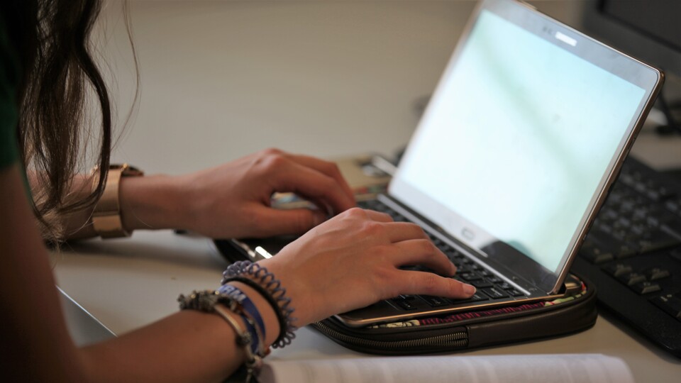 Die Hände einer Frau tippen auf einem Laptop. Daneben liegt ein aufgeschlagenes Buch.