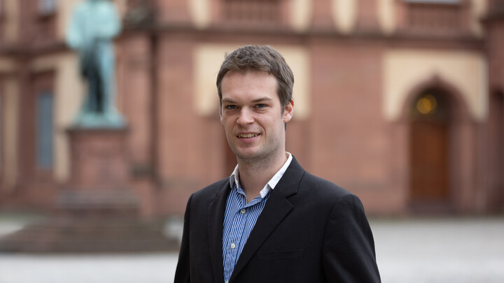 Marius Krömer. He is wearing a black suit and is standing in front of the castle on the Ehrenhof.
