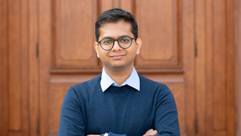 Tanay Agarwal. He is wearing a blue sweater over a white shirt and is standing in front of brown door.