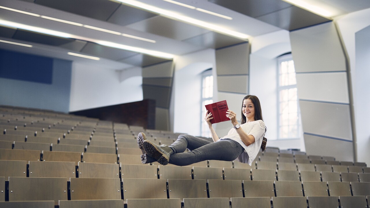 Studentin mit weißer Bluse und grauer Hose liegt mit den Beinen überschränkt und einem Buch mit der Aufschrift "Deutsche Gesetzte" in der Hand schwebend in der Luft über den Stüheln eines Hörsaals.