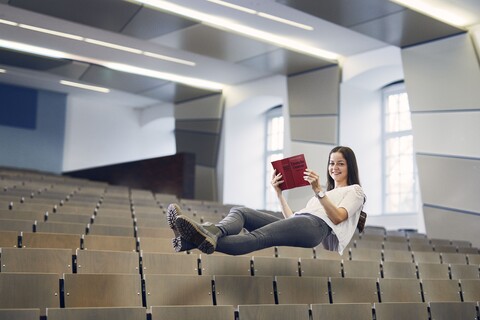 Studentin mit weißer Bluse und grauer Hose liegt mit den Beinen überschränkt und einem Buch mit der Aufschrift "Deutsche Gesetzte" in der Hand schwebend in der Luft über den Stüheln eines Hörsaals.