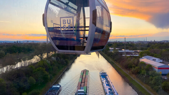 Eine Gondel mit der Aufschrift 'BUGA' schwebt über dem Neckar bei Sonnenuntergang, darunter sind drei Frachtschiffe zu sehen.