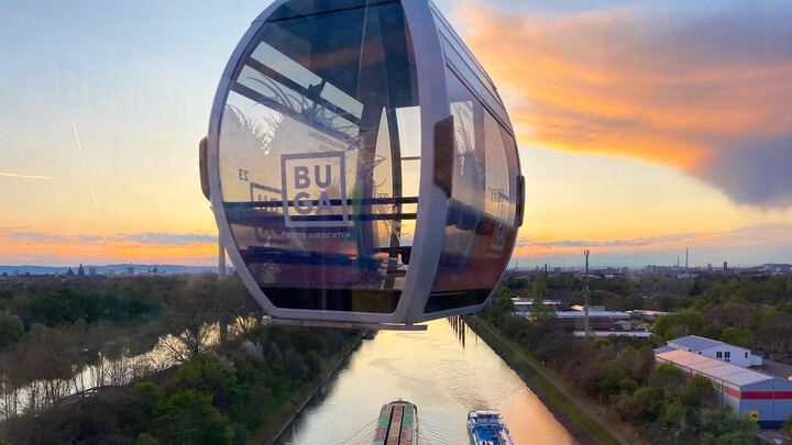 Eine Gondel mit der Aufschrift 'BUGA' schwebt über dem Neckar bei Sonnenuntergang, darunter sind drei Frachtschiffe zu sehen.