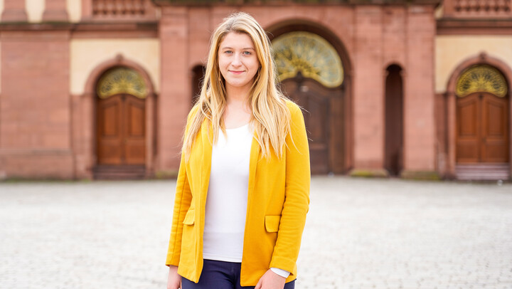 Eine Person trägt ein weißes T-Shirt sowie einen gelben Blazer und steht vor dem Schloss der Universität Mannheim. Die Person heißt Monika Buknova.