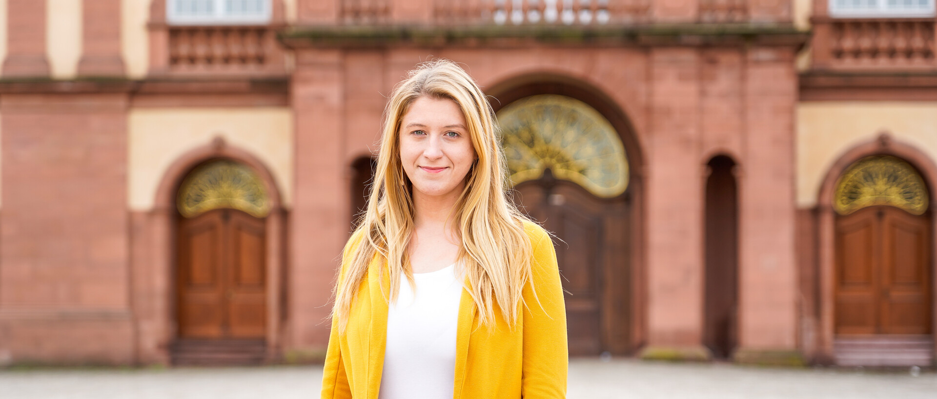 Eine Person trägt ein weißes T-Shirt sowie einen gelben Blazer und steht vor dem Schloss der Universität Mannheim. Die Person heißt Monika Buknova.