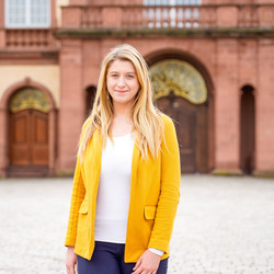 Eine Person trägt ein weißes T-Shirt sowie einen gelben Blazer und steht vor dem Schloss der Universität Mannheim. Die Person heißt Monika Buknova.
