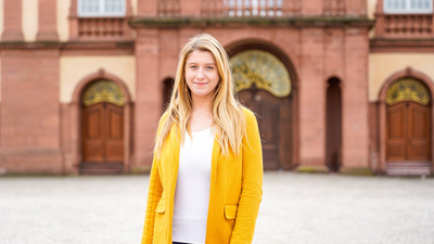 Eine Person trägt ein weißes T-Shirt sowie einen gelben Blazer und steht vor dem Schloss der Universität Mannheim. Die Person heißt Monika Buknova.