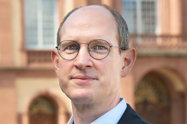 Herr Spalt trägt ein dunkles Jacket. Er hat eine Brille. Im Hintergrund ist eien Schlossfassade zu sehen.