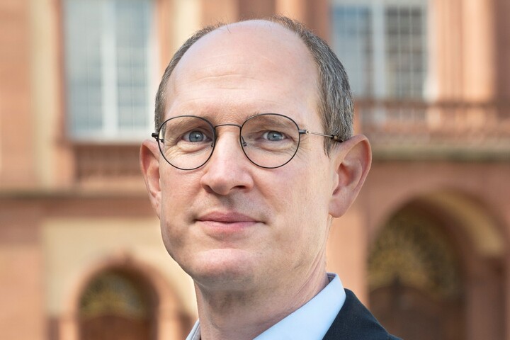 Herr Spalt trägt ein dunkles Jacket. Er hat eine Brille. Im Hintergrund ist eien Schlossfassade zu sehen.