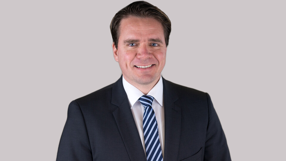 Hartmut Höhle. He is wearing a black suit with a stripped tie in front of a white background.