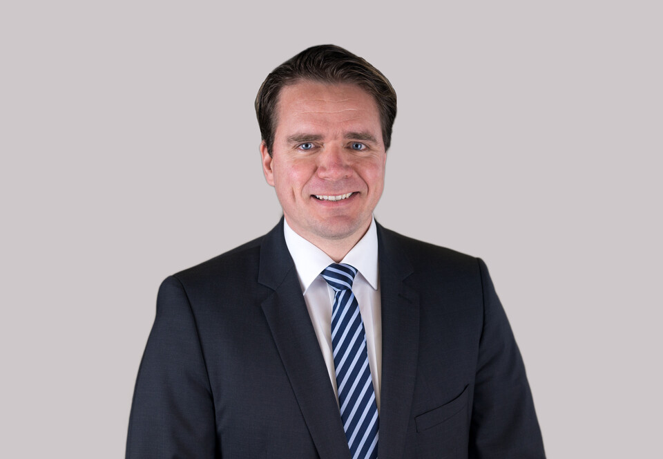 Hartmut Höhle. He is wearing a black suit with a stripped tie in front of a white background.