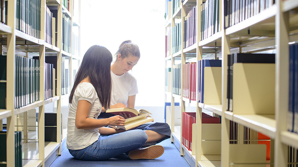 Zwei Studentinnen sitzen in der Bibliothek auf dem Boden vor dem Regel und blättern in Büchern