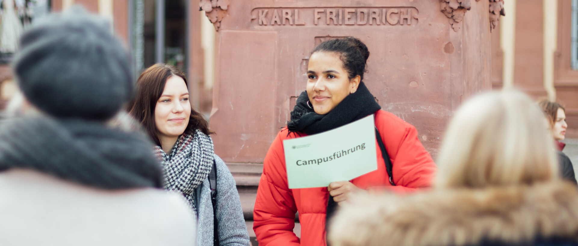 Umringt von Studierenden hält eine Campusführerin vor dem Denkmal Karl Friedrichs auf dem Ehrenhof ein Schild mit der Aufschrift Campusführung