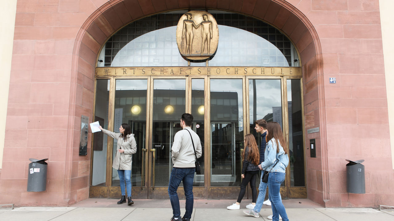 Studierende bei einer Führung vor dem Haupteingang mit der Aufschrift "Wirtschaftshochschule"